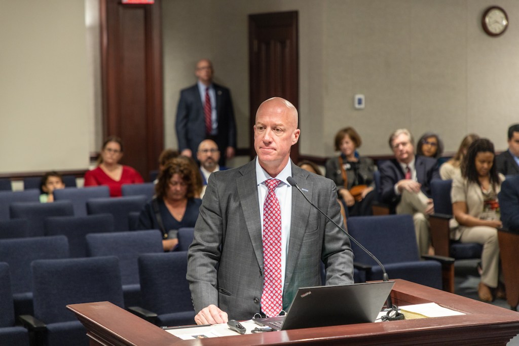 A bald man in a gray suit and a red patterned tie is speaking at a wooden podium. He is in a large room with rows of dark blue seats, some of which are occupied by an audience in the background.