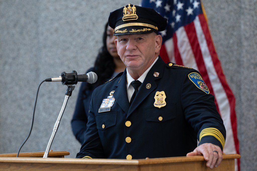 A man in a police uniform stands at a podium. An American flag is behind him.