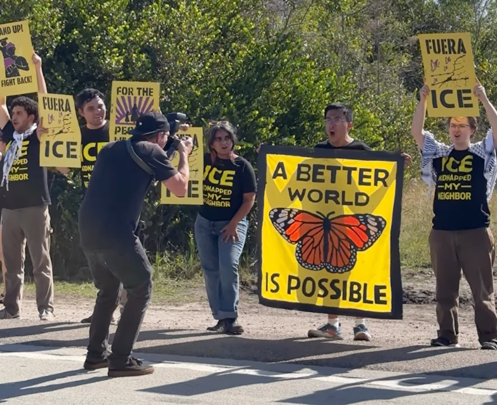A group of protestors stands outside holding yellow and black signs. A cameraman films a man holding a large yellow banner featuring a monarch butterfly and the text, "A BETTER WORLD IS POSSIBLE."